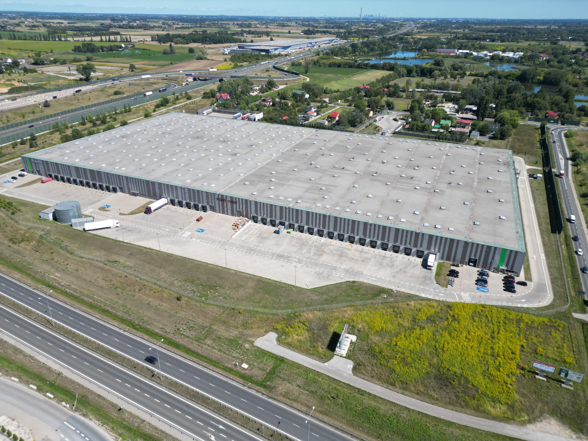 Szeroki kadr z lotu ptaka ukazuje rozległy magazyn z rampami załadunkowymi, otoczony zielenią i drogami dojazdowymi. W tle widoczna zabudowa miejska.