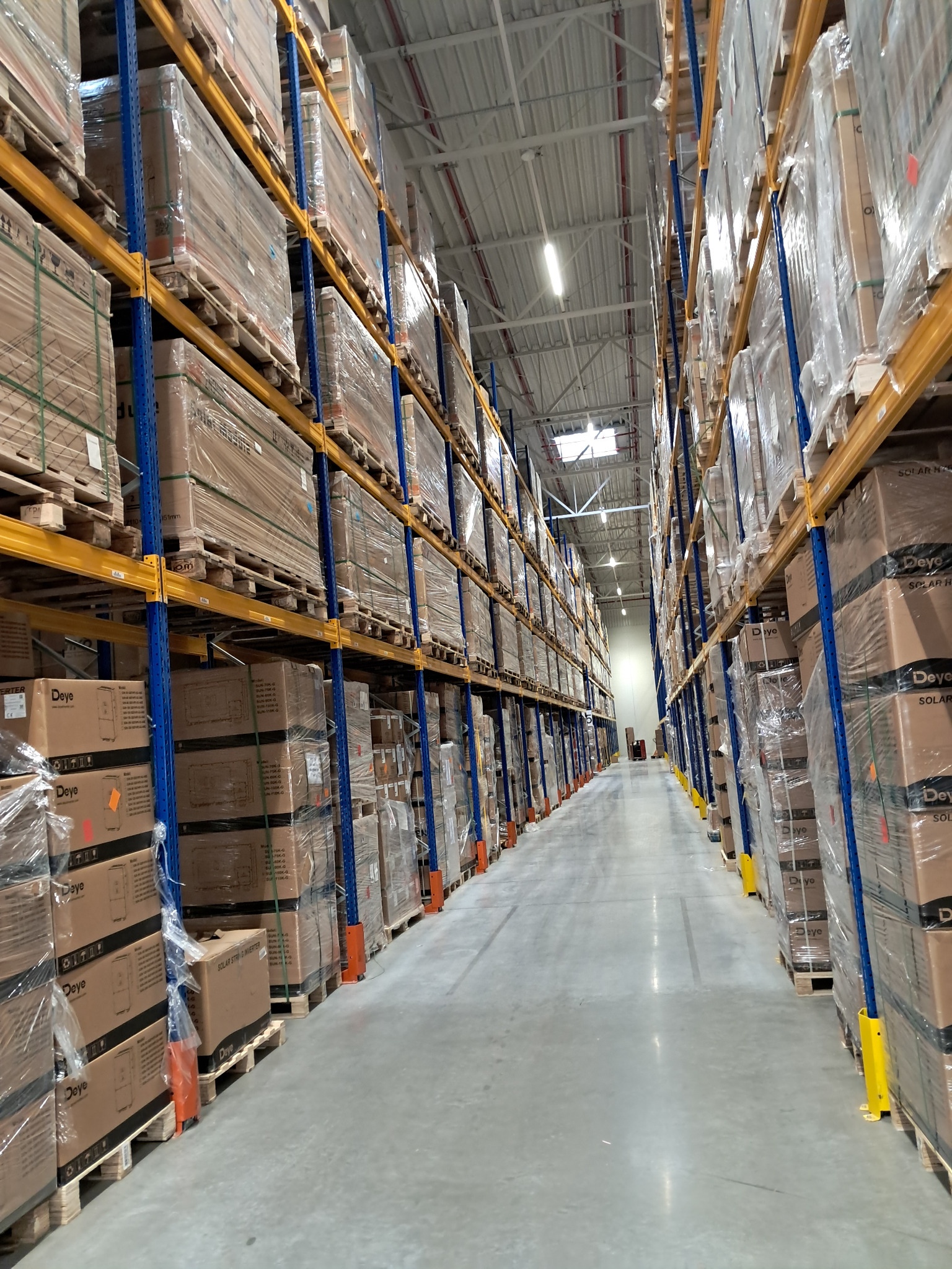 Wnętrze magazynu z wysokimi regałami wypełnionymi kartonami na paletach, zabezpieczonymi folią. Widok na długi korytarz między regałami. Metalowa konstrukcja regałów.