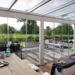 Rafał Leoniewski Solutions - Zadaszony taras z przesuwnymi szklanymi ścianami, drewnianą podłogą i widokiem na ogród z elementami małej architektury, takimi jak donice i ławka.