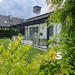 Rafał Leoniewski Solutions - Szklana zabudowa tarasu z czarną ramą, widok z ogrodu przez zielone liście. Wewnątrz tarasu rattanowe meble ogrodowe, na dachu panele słoneczne. W rogu widoczne logo firmy.