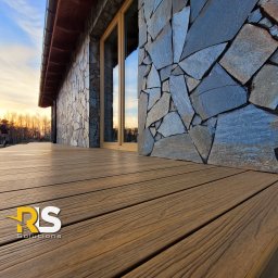 Rafał Leoniewski Solutions - Nowoczesny taras z desek kompozytowych w odcieniach brązu, przylegający do ściany budynku wykończonej kamieniem naturalnym w różnych odcieniach szarości, okno z drewnianą ramą, widok na zachodzące...