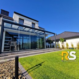 Rafał Leoniewski Solutions - Nowoczesna, przeszklona konstrukcja tarasowa z czarnymi ramami, przylegająca do białego domu, widoczna z poziomu zadbanego trawnika w słoneczny dzień.