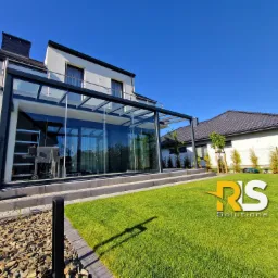 Nowoczesna, przeszklona konstrukcja tarasowa z czarnymi ramami, przylegająca do białego domu, widoczna z poziomu zadbanego trawnika w słoneczny dzień.