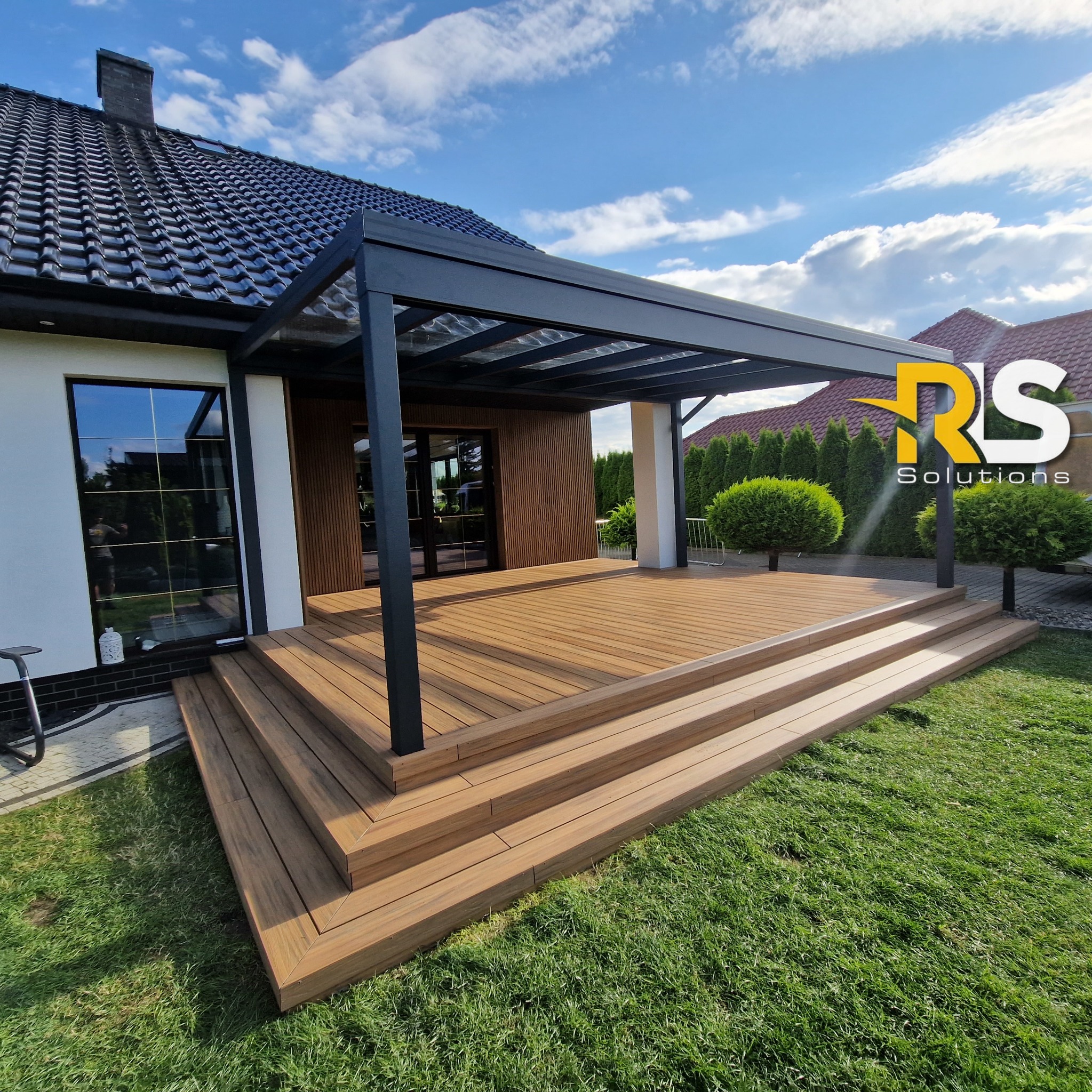 Nowoczesny taras z desek kompozytowych z czarną metalową pergolą przylegający do domu z drewnianą elewacją, widok z poziomu trawnika w słoneczny dzień.