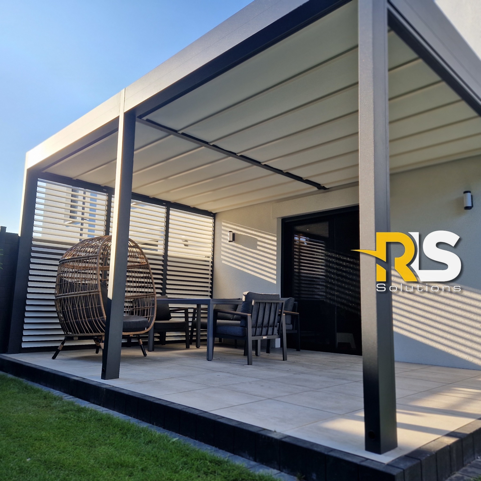 Nowoczesna pergola z metalową konstrukcją i zadaszeniem lamelowym, uzupełniona o rattanowy fotel wiszący i zestaw mebli ogrodowych na płytkach tarasowych.