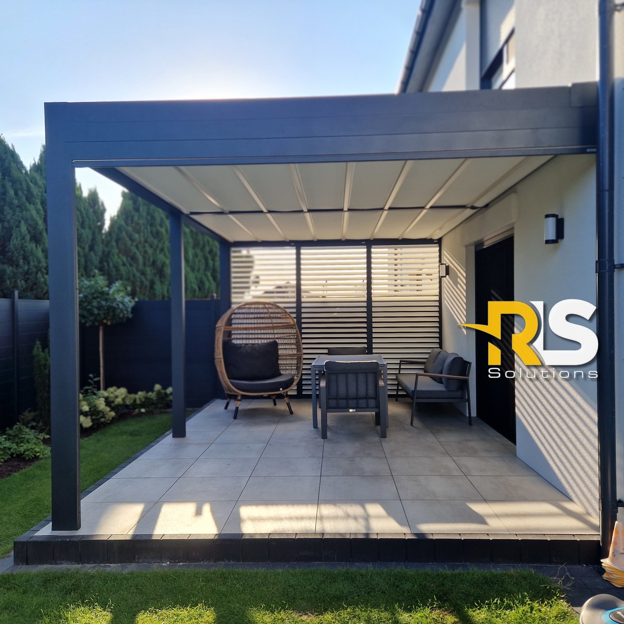 Nowoczesna, antracytowa pergola metalowa z regulowanym zadaszeniem, wyposażona w rattanowy fotel i zestaw mebli ogrodowych, przylegająca do jasnej elewacji domu z widocznym logo firmy.