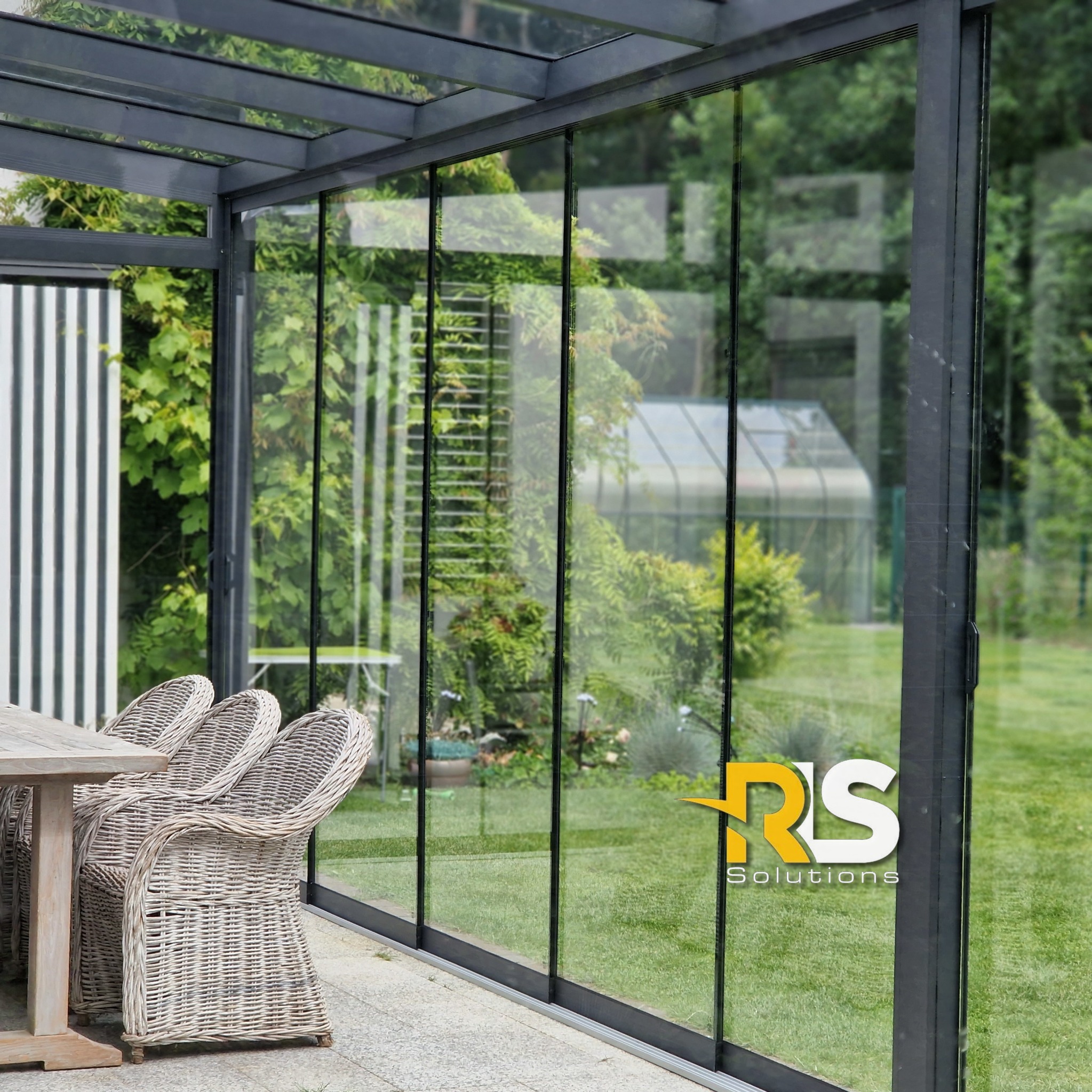 Zadaszony taras z przesuwnymi szklanymi panelami, widoczne wiklinowe krzesła i stół, w tle zieleń ogrodu i szklarnia.