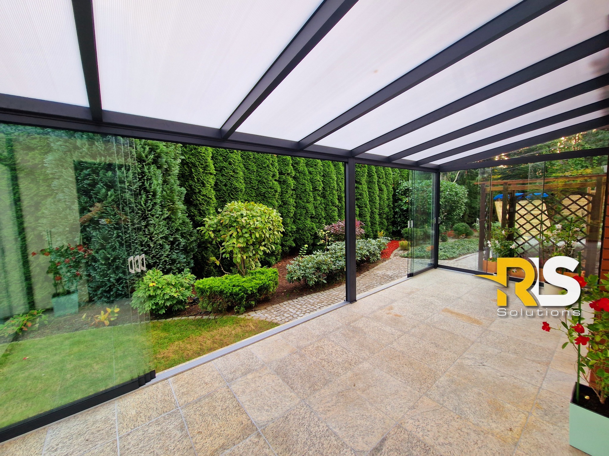 Zadaszony taras z przeszklonymi ścianami i metalową konstrukcją, widok na zadbany ogród z tujami i kwitnącymi roślinami, jasna kamienna posadzka.