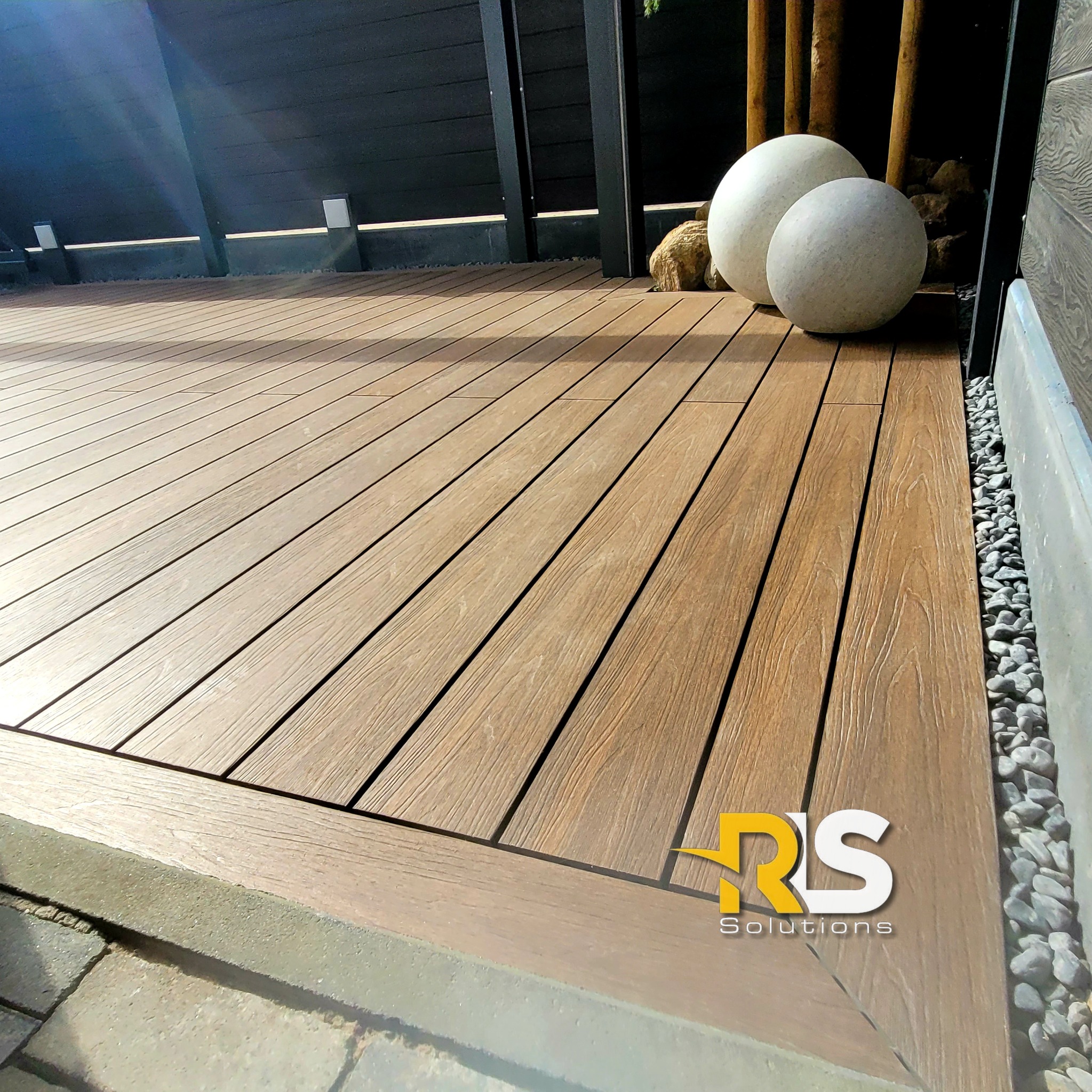 Nowoczesny taras z desek kompozytowych w kolorze drewna, z szarymi kulami dekoracyjnymi i czarnym ogrodzeniem w tle, widoczne logo firmy w prawym dolnym rogu.