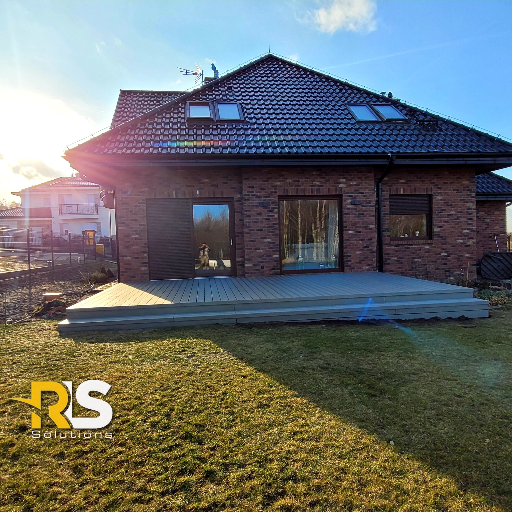 Szary taras kompozytowy z trzema stopniami, przylegający do ceglanego domu z czarnym dachem, widok z trawnika w słoneczny dzień, z logo firmy w rogu.