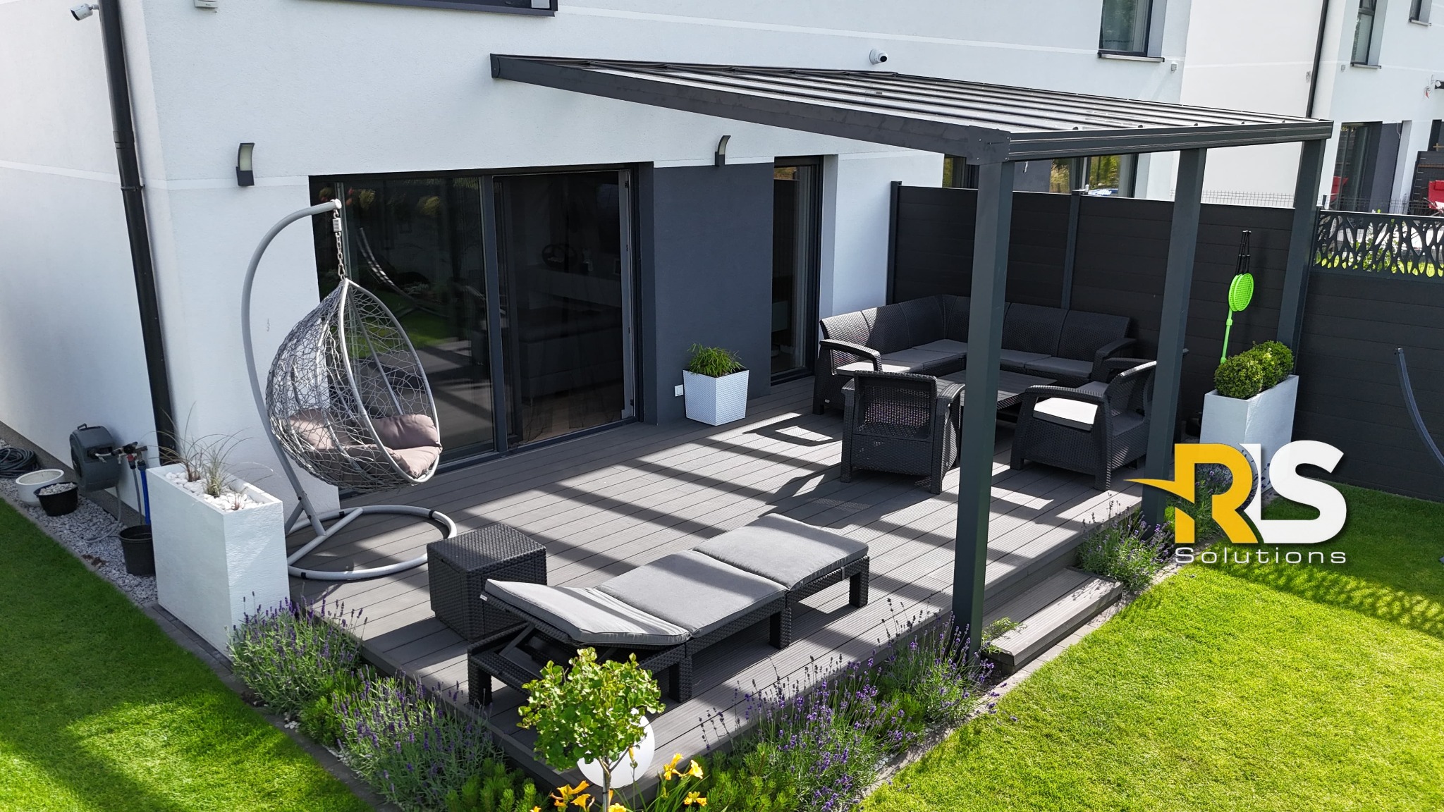 Nowoczesny taras z szarej deski kompozytowej, zadaszony metalową pergolą, wyposażony w meble ogrodowe z rattanu i wiszące krzesło, otoczony zielonym trawnikiem i rabatami lawendy.
