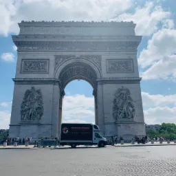 Bus firmy Uniwer-Trans zaparkowany przed Łukiem Triumfalnym w Paryżu, błękitne niebo z chmurami w tle.