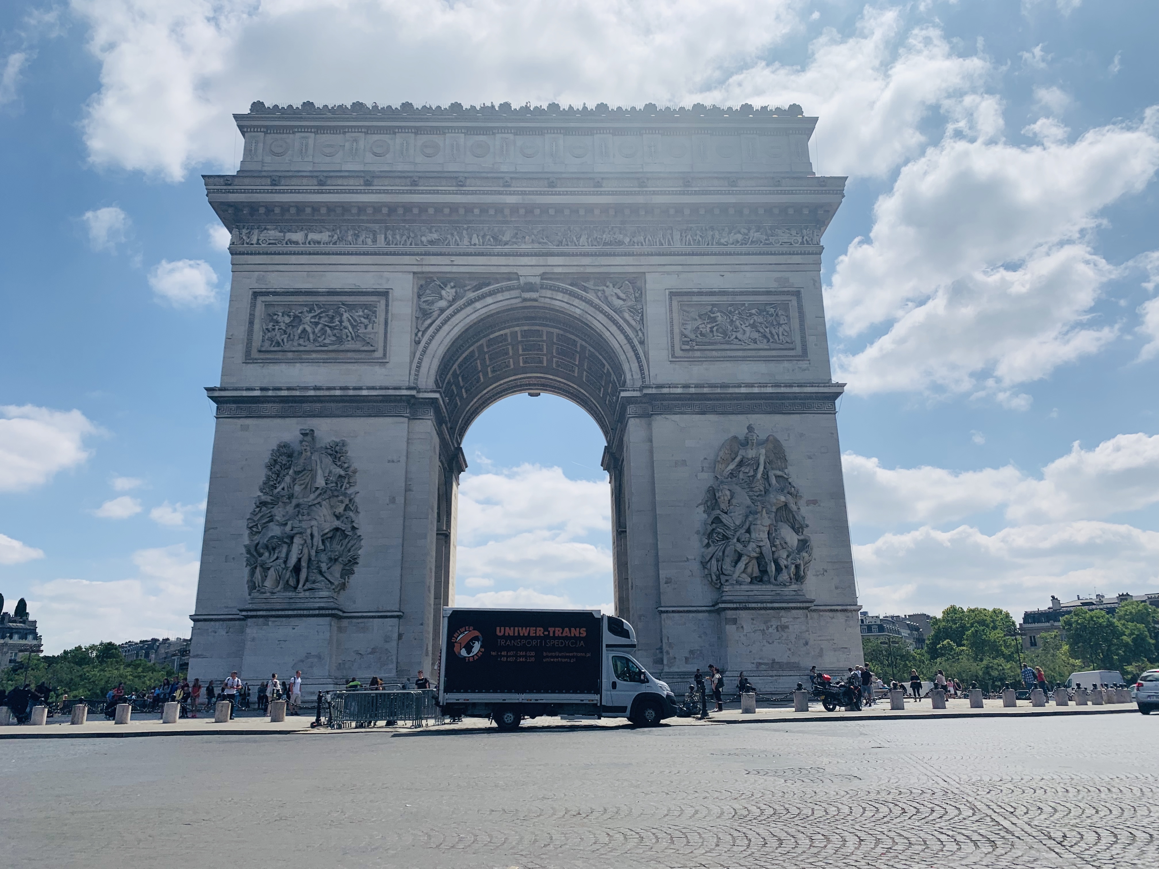 Bus firmy Uniwer-Trans zaparkowany przed Łukiem Triumfalnym w Paryżu, błękitne niebo z chmurami w tle.