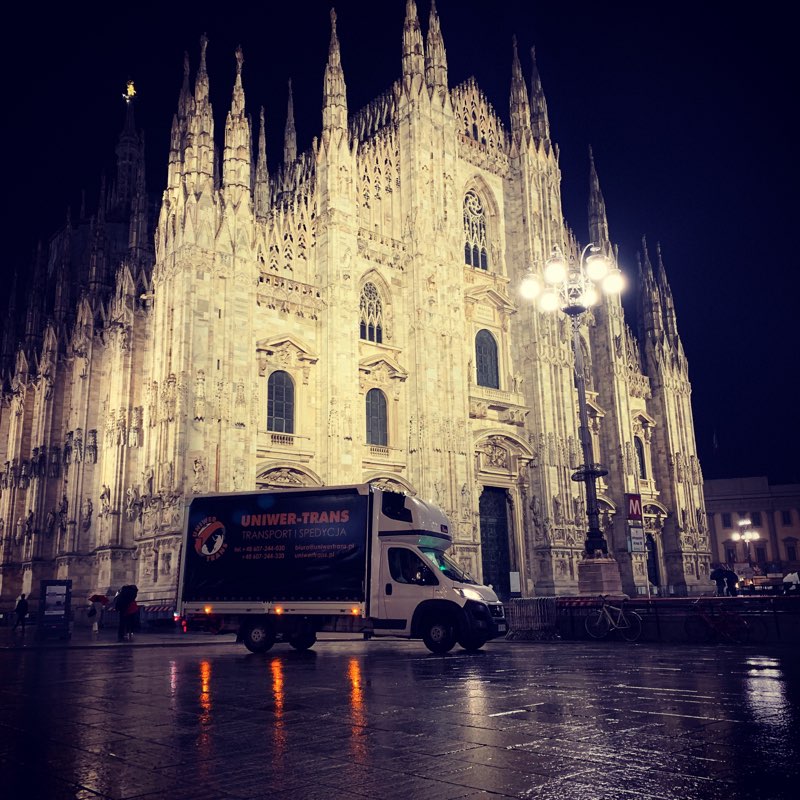Biały bus firmy transportowej Uniwer-Trans na tle oświetlonej Katedry w Mediolanie, nocna sceneria z mokrym placem odbijającym światła.