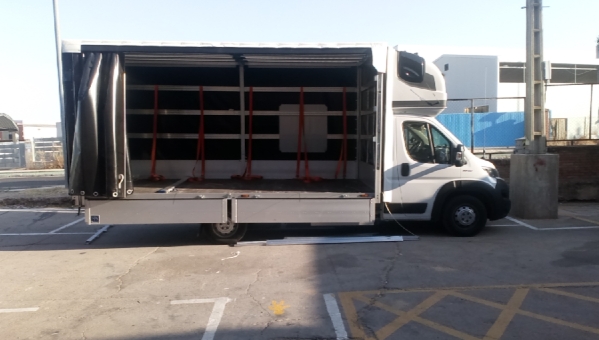 Biały bus dostawczy z otwartą tylną przestrzenią ładunkową, zabezpieczoną czarną plandeką częściowo odsłoniętą, puste wnętrze z zabezpieczeniami do transportu delikatnych przedmiotów, na parkingu.