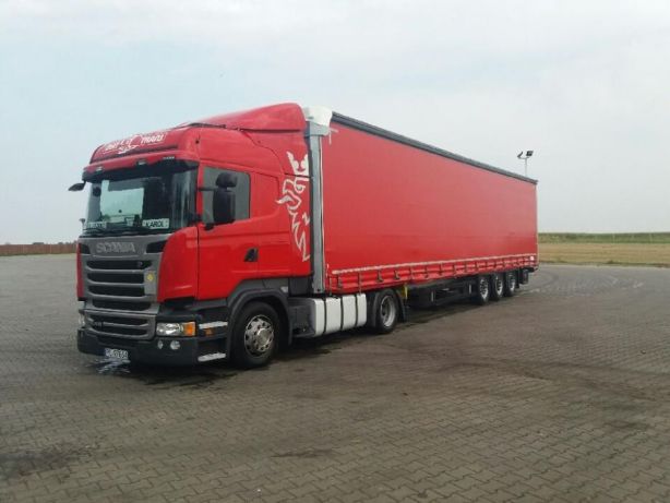 Czerwona ciężarówka Scania z naczepą na brukowanym placu, widoczne logo firmy transportowej na kabinie.