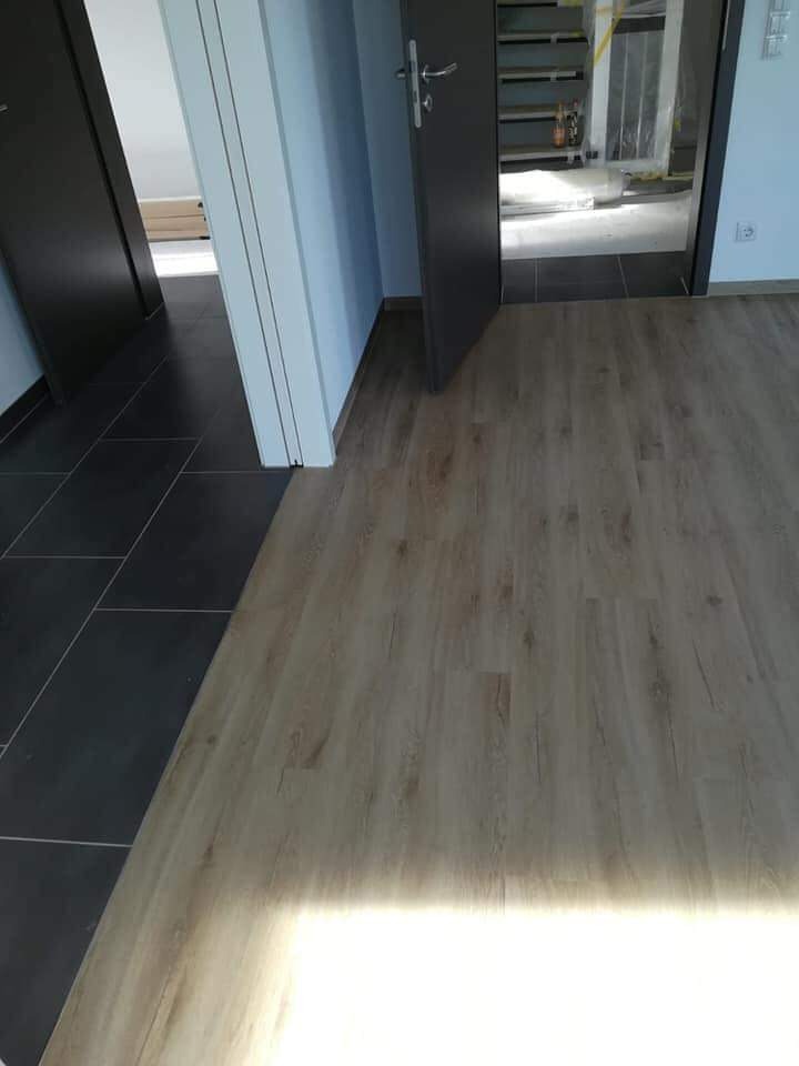 Ułożone panele podłogowe w jasnym odcieniu drewna, kontrastujące z ciemnymi płytkami w sąsiednim pomieszczeniu, widok na otwarte drzwi i wnętrze w trakcie prac wykończeniowych.