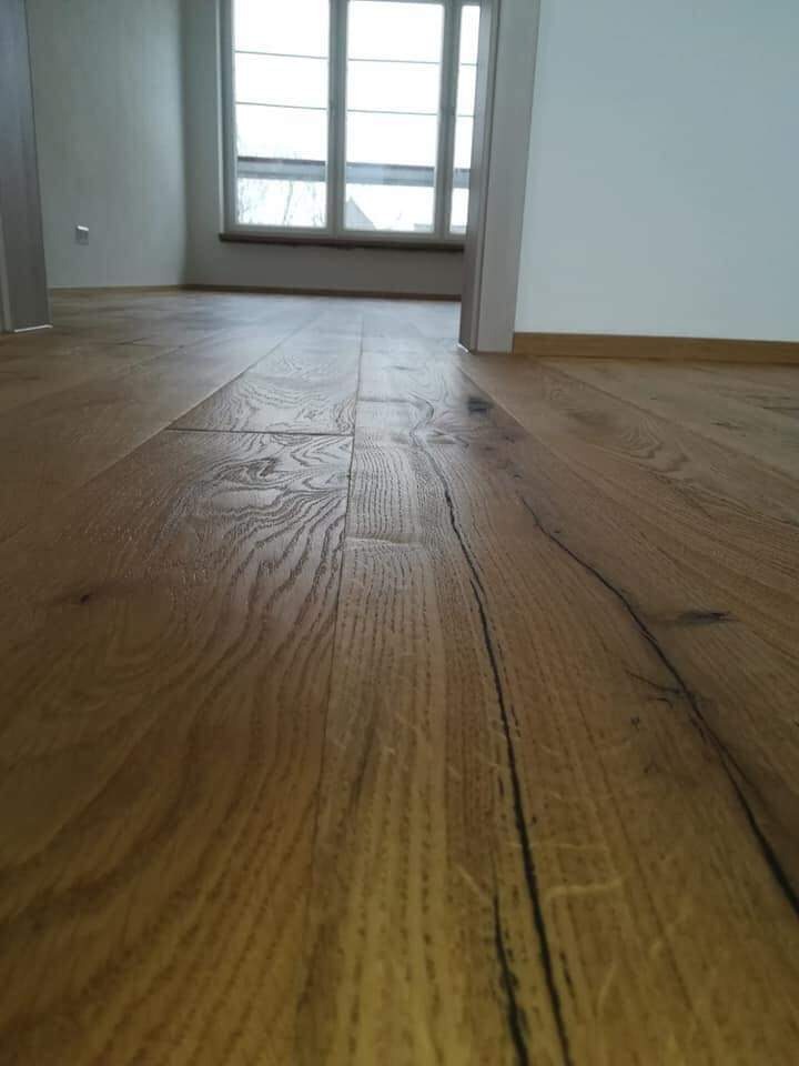 Ułożony parkiet z wyraźnym usłojeniem, widoczne sęki i pęknięcia, w tle okno z białymi ramami i fragment białej ściany, perspektywa z niskiego kąta.