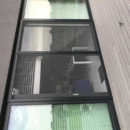 Cztery okna z szarymi ramami, z których jedno ma zamontowaną moskitierę, widok z zewnątrz na ścianę z szarym tynkiem i elewację z jasnych paneli.