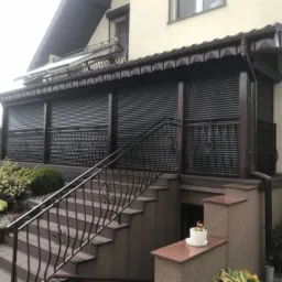 Zewnętrzny taras z zabudowanymi, ciemnymi roletami, widok z dołu schodów z ozdobną balustradą, mała doniczka z kwiatami na murku.