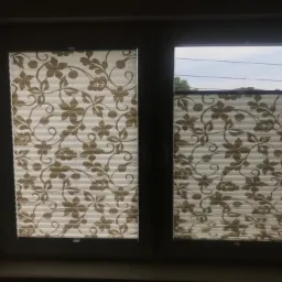 Dwuokienne okno z plisowanymi, beżowo-białymi roletami wewnętrznymi w kwiatowy wzór, częściowo zasłonięte, z widokiem na zewnątrz przez niezaciemnioną część okna.