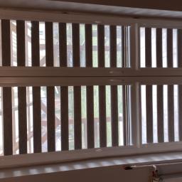 RolloTrans Paweł Kaźmierczak - Zamknięte, pionowe żaluzje w oknie z białymi ramami, widok na zieleń za oknem, grzejnik pod parapetem.