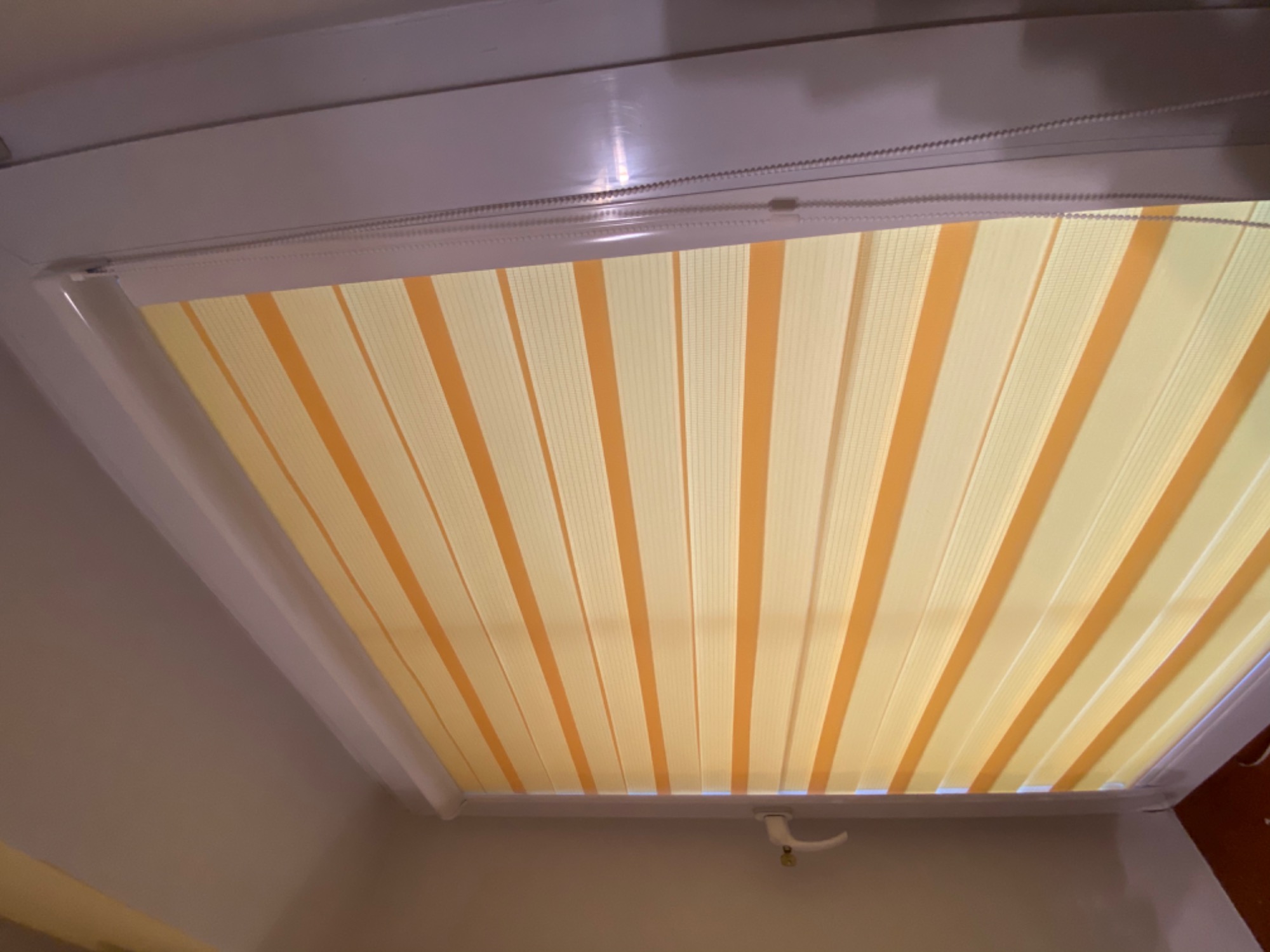 Żółto-białe, pionowe pasy żaluzji okiennej w jasnej ramie, widok od dołu, uchwyt do regulacji widoczny w prawym dolnym rogu.