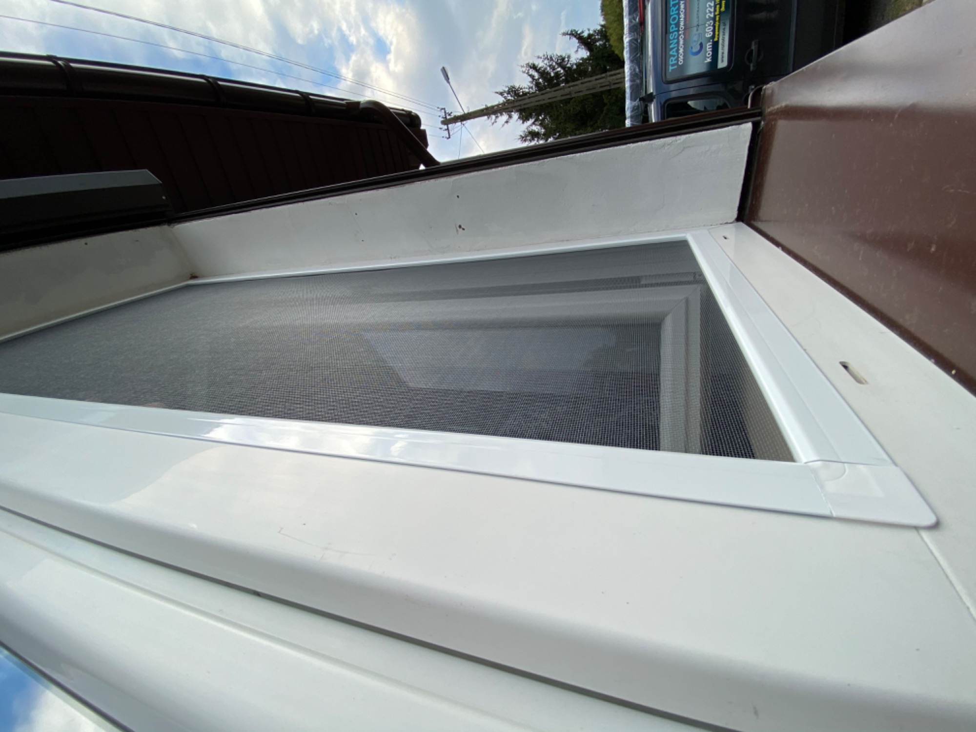 Zamontowana moskitiera w oknie z białymi ramami, widoczna z bliskiej perspektywy od dołu, z fragmentem dachu i samochodu w tle.