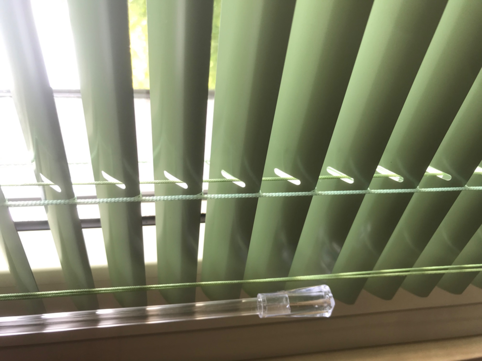 Fragment pionowych, oliwkowych żaluzji z widocznym sznurkiem i przezroczystym uchwytem do regulacji, z jasnym światłem wpadającym przez okno.