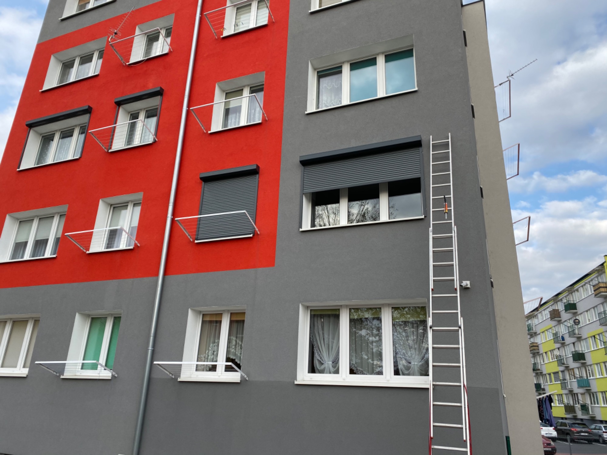 Fasada budynku z zamontowanymi roletami zewnętrznymi, widoczna drabina oparta o ścianę. Część ściany pomalowana na intensywny czerwony kolor, pozostała część w odcieniach szarości.