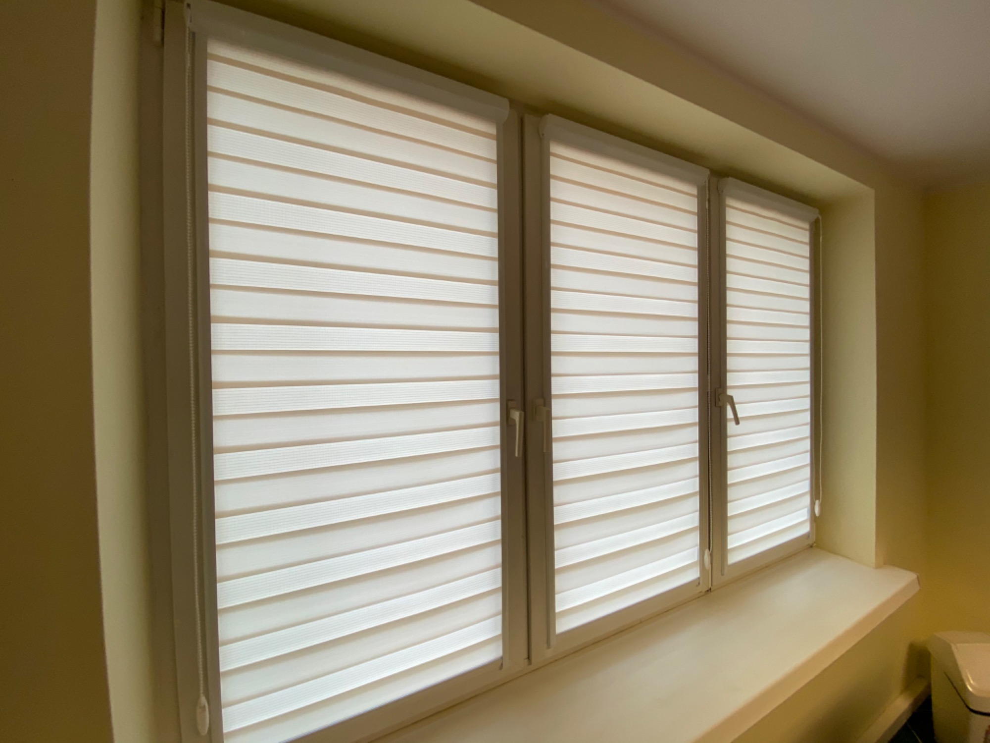 Trzy okna z zamontowanymi roletami dzień i noc w kolorze kremowym, częściowo zasłonięte, widoczne sznurki sterujące.