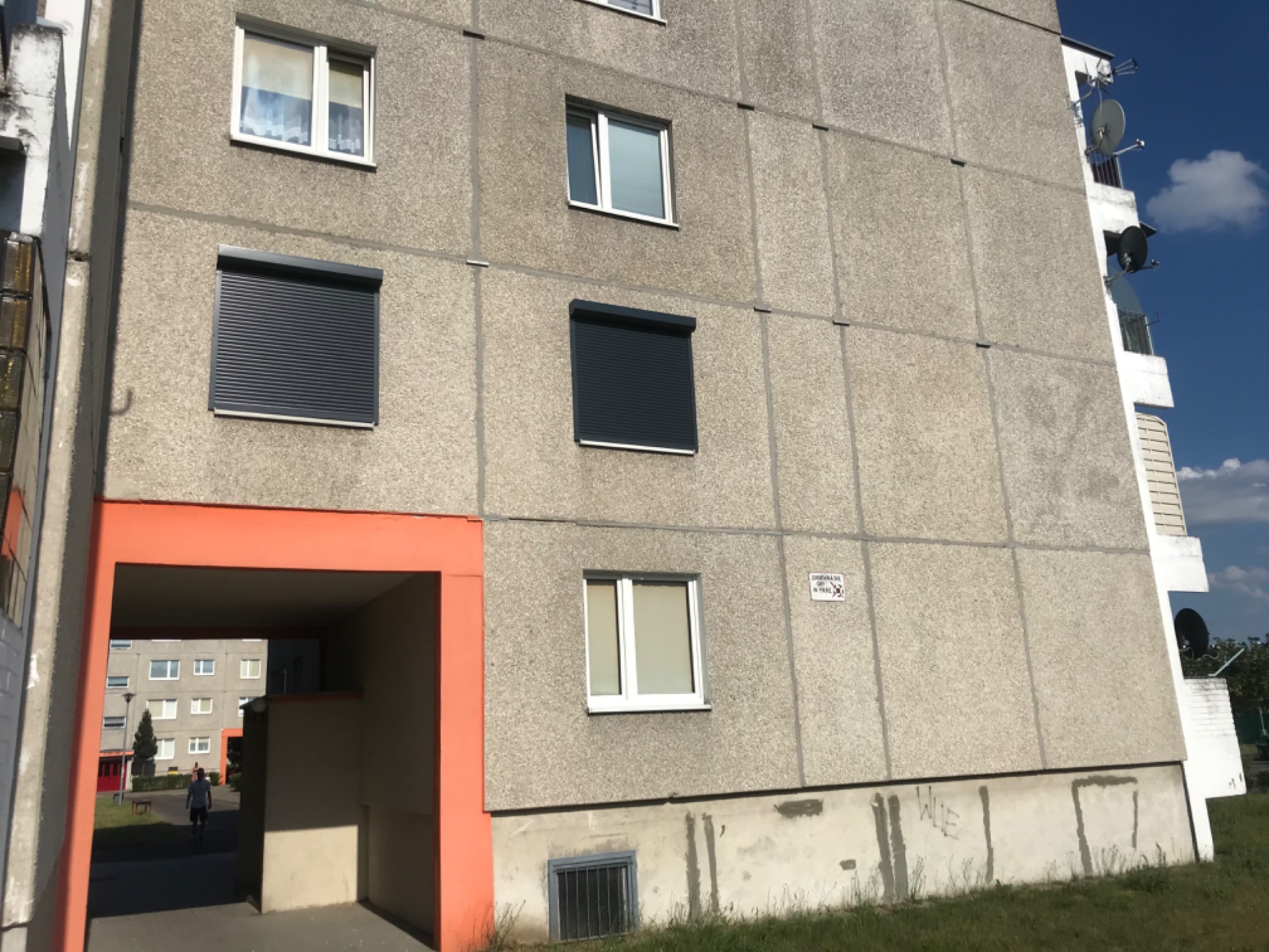 Szary blok mieszkalny z dwoma oknami zabezpieczonymi opuszczonymi roletami zewnętrznymi, pozostałe okna bez rolet, widok z dołu, widoczny fragment przejścia w kolorze pomarańczowym.