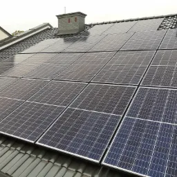 Czarny dach pokryty panelami słonecznymi, widoczny komin z cegły na tle pochmurnego nieba.