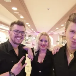 Trzyosobowa grupa w czarnych strojach pozuje na tle sali bankietowej z dekoracjami kwiatowymi i oświetleniem sufitowym.