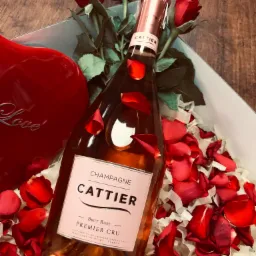 Butelka różowego szampana Cattier Premier Cru w pudełku prezentowym z czerwoną dekoracją w kształcie serca z napisem 'Love' i płatkami róż, otoczona świeżymi czerwonymi różami.
