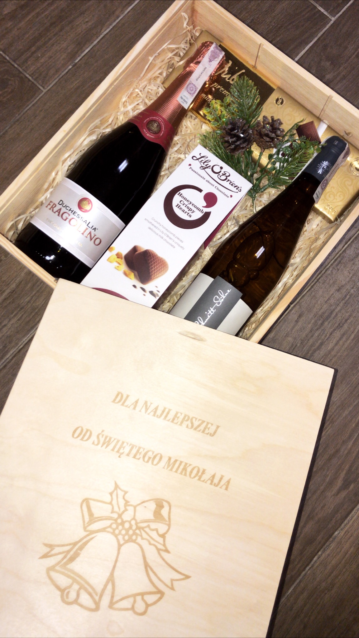 Świąteczny kosz prezentowy z Łochowa: dwie butelki wina, czekoladki Lily O'Brien's i dekoracje z gałązek świerku oraz szyszek w drewnianej skrzynce z grawerem 'Dla Najlepszej od Świętego Mikołaja'...