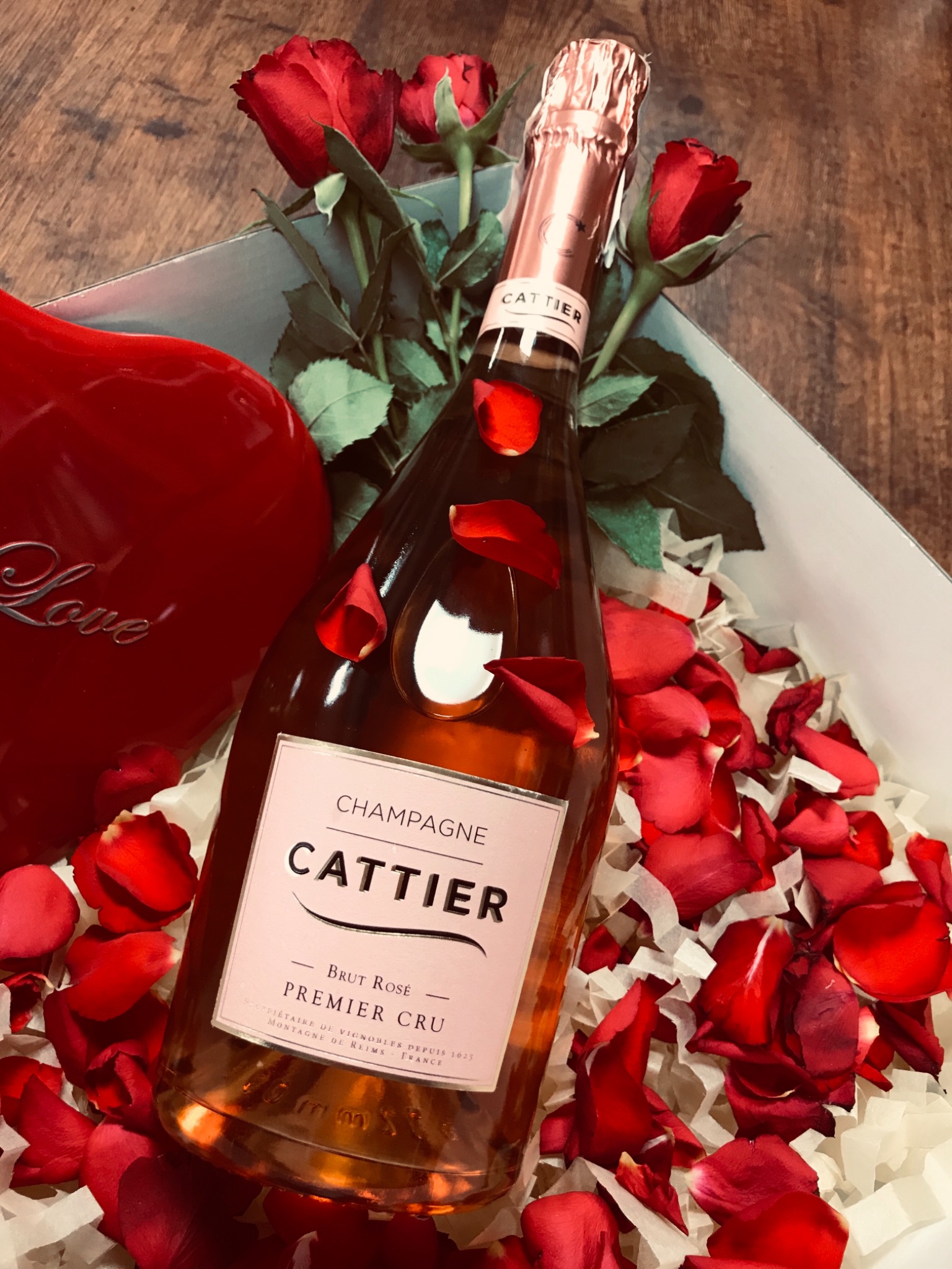Butelka różowego szampana Cattier Premier Cru w pudełku prezentowym z czerwoną dekoracją w kształcie serca z napisem 'Love' i płatkami róż, otoczona świeżymi czerwonymi różami.