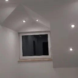 Świeżo otynkowane, gładkie ściany poddasza ze skosami i oknem, oświetlone punktowymi lampami sufitowymi.