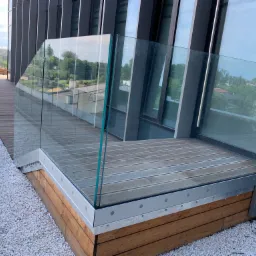 Nowoczesny balkon z drewnianą podłogą i szklaną balustradą, widok z zewnątrz, otoczony białym żwirem i elementami architektury miejskiej w tle.