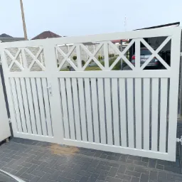 Nowoczesna, biała brama wjazdowa z pionowymi szczeblami i dekoracyjnym wzorem krzyżowym w górnej części, na tle zadbanego podwórka z kostki brukowej i widocznym samochodem w tle.