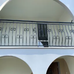 Czarna, metalowa balustrada balkonowa z dekoracyjnymi elementami w kształcie litery C, zamontowana na balkonie z łukowym wejściem w budynku o jasnej elewacji.