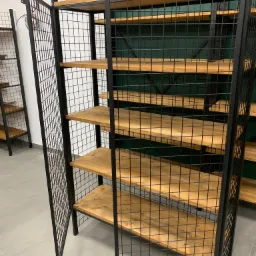 Metalowa szafka z drewnianymi półkami i otwieranymi drzwiczkami z siatki, widoczne łączenia spawane i nitowane, w tle zielona ściana.