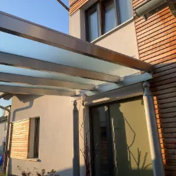 Zadaszenie tarasu z przezroczystego tworzywa sztucznego, wsparte na metalowych słupach, przylegające do budynku z fasadą wykończoną drewnem i jasnym tynkiem, widoczne okno z drewnianymi żaluzjami.