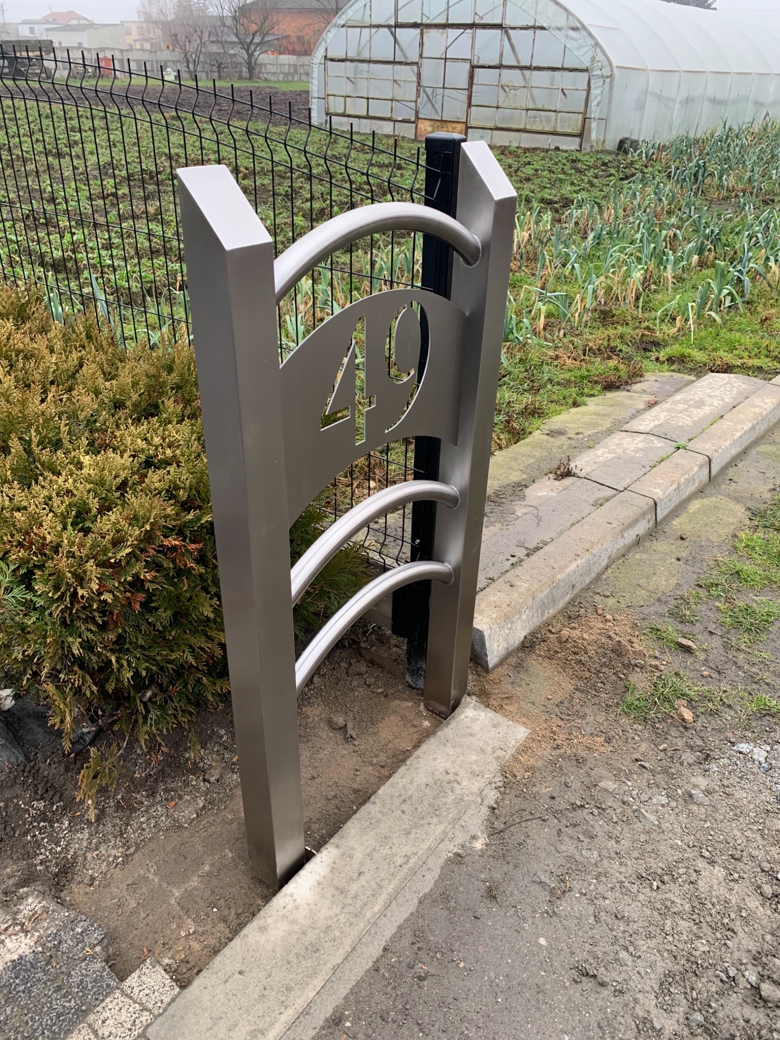 Metalowa tabliczka z numerem 49 na tle siatki ogrodzeniowej i szklarni, świeżo zamontowana w ziemi.