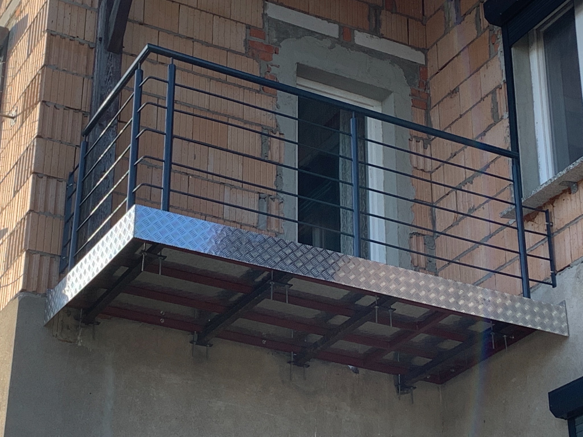 Balkon z metalową podłogą w romby i czarną, minimalistyczną balustradą, zamontowany przy budynku z cegły dziurawki w trakcie budowy.