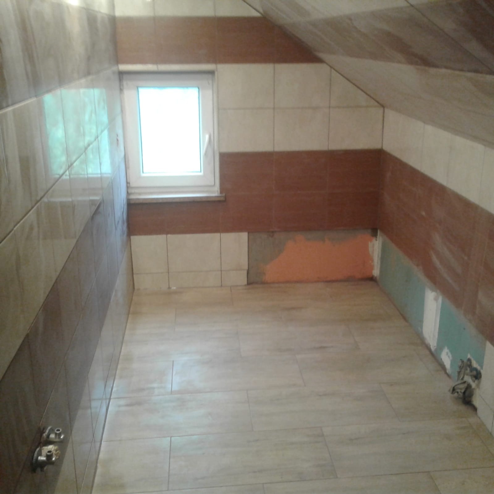 Łazienka w trakcie remontu: częściowo ułożone brązowo-beżowe płytki na ścianach i podłodze, widoczne fragmenty surowej ściany z resztkami farby i kleju, małe okno z białymi ramami.