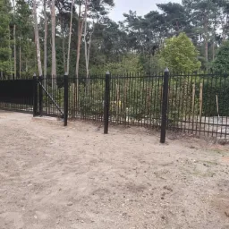 Czarna metalowa brama wjazdowa z pionowymi prętami i szpicami, widoczna na tle lasu, z piaskowym podłożem, częściowo zamontowana.