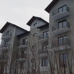 Surowy budynek wielorodzinny w trakcie wykończenia, z zamontowanymi czarnymi balustradami balkonowymi o poziomym układzie szczebli, widziany z dołu przez gałęzie drzew bez liści.