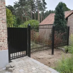 Czarna, metalowa furtka i przęsło ogrodzenia z pionowymi szczebelkami, zakończonymi ostrymi grotami, na tle ceglanego domu i zieleni.