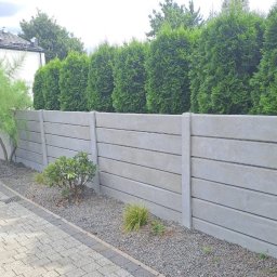 Wulkabet Producent Ogrodzeń Betonowych - Szary, poziomy płot betonowy z roślinami i drzewami w tle, widziany z niskiej perspektywy. Płot oddziela posesję od zieleni, tworząc estetyczne ogrodzenie.
